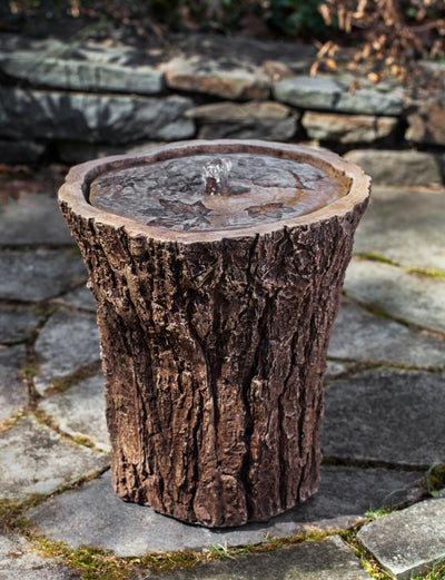 A cylindrical fountain that looks like it's carved out of wood, with a rough bark exterior and leaves imprinted in the top plate of the fountain.