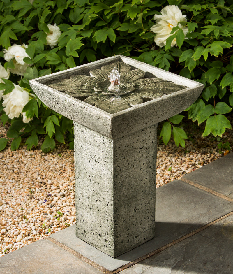 A square fountain on a square pedestal, with a flower design on the top. Pictured in front of white flowers.