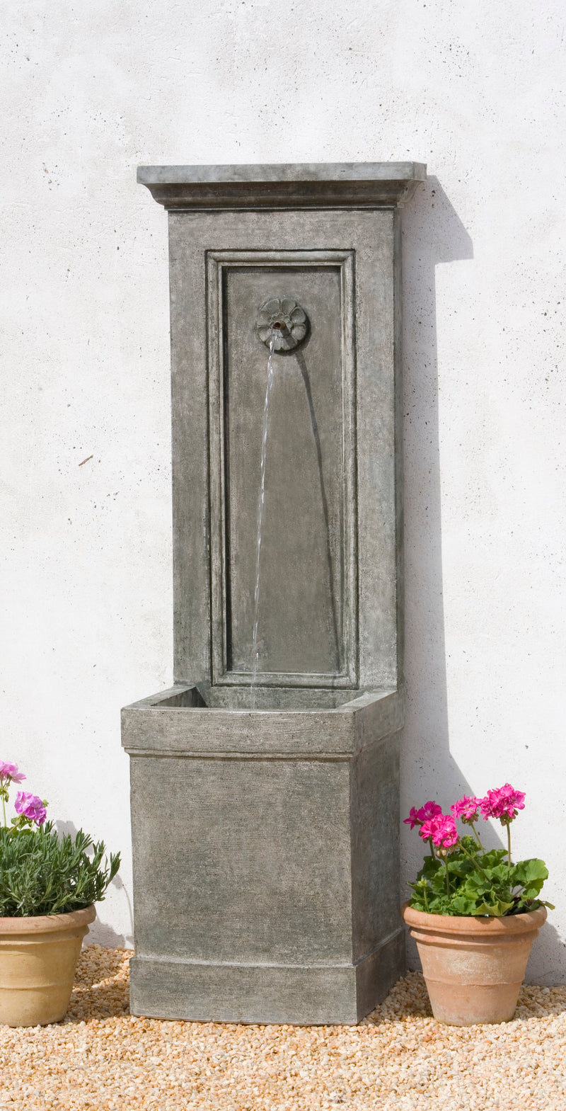 Tall, narrow, grey fountain placed against a white wall, flanked by planted terra cotta planters and pink flowers