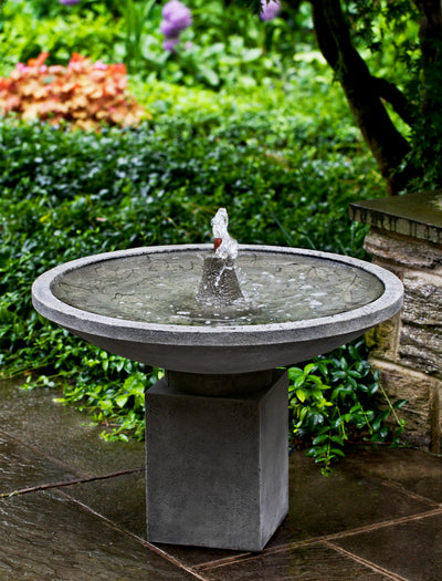 A round bowl with a spout on top of a rectangular pedestal, pictured on hardscape and in front of foliage
