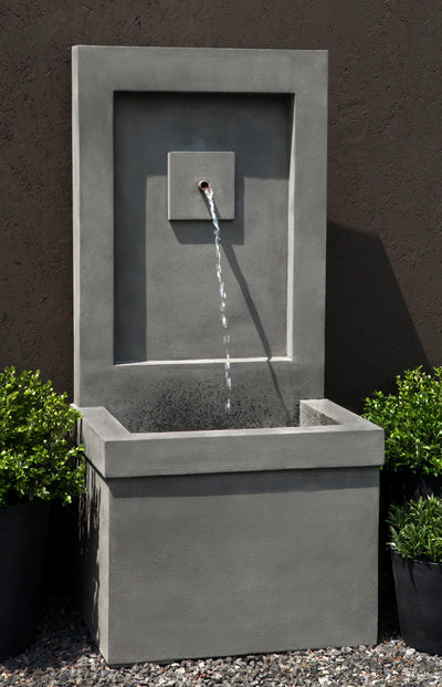 Tall square grey fountain with a single spout, flanked by green shrubs