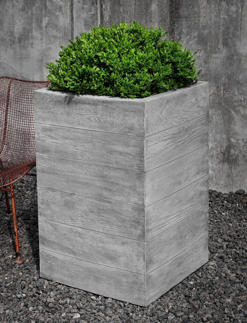 Tall square cast stone container planted with a boxwood and shown next to a metal chair