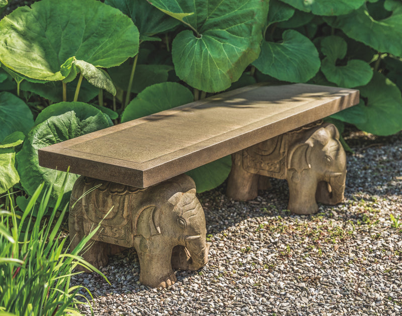 Straight, brown bench with stylized elephants as legs, pictured in front of large green leaves