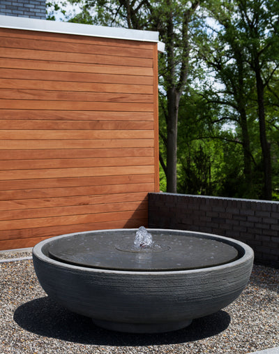 Large bowl shaped fountain with covered top, pictured on gravel in front of wood pannel