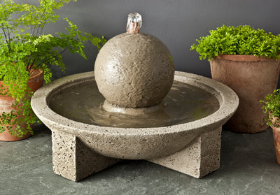 Tabletop fountain with a center sphere in a basin pictured in front of planted ferns