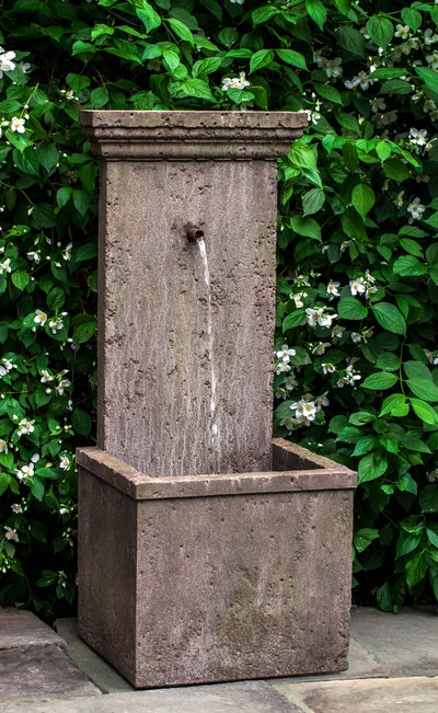 Wall fountain with one spout dropping into a square basin, shown in front of greenery