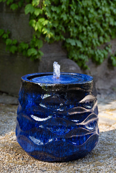 Short faceted blue fountain with small bubbler pictured in front a ivy-covered wall