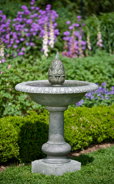 Gray fountain with pineapple finial pictured in front of shrubs