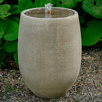 Small grey fountain in vertical oval shape with copper spout in front of green leaves and sitting on gravel 