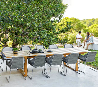 Rectangular teak table with ten chairs shown on patio with three people talking on the side