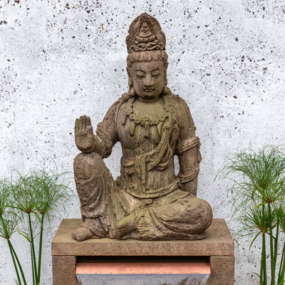 An ornate Buddha statue rests on top of a square fountain basin, with water flowing over a copper water spiller. Flanked by potted papyrus plants.