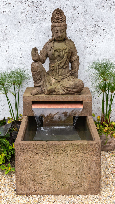 An ornate Buddha statue rests on top of a square fountain basin, with water flowing over a copper water spiller. Flanked by potted papyrus plants.