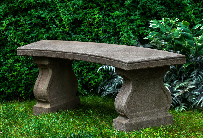 Curved cast stone bench pictured in front of green foliage