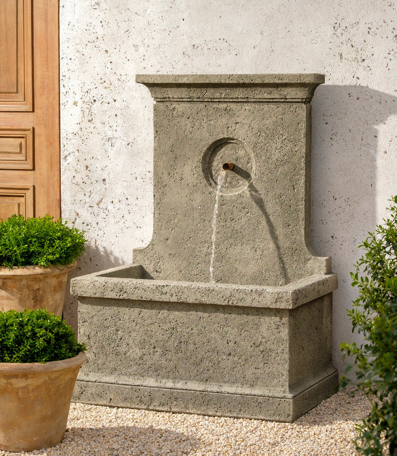 Tall, narrow fountain against a white wall, flanked by terracotta planters planted with boxwood