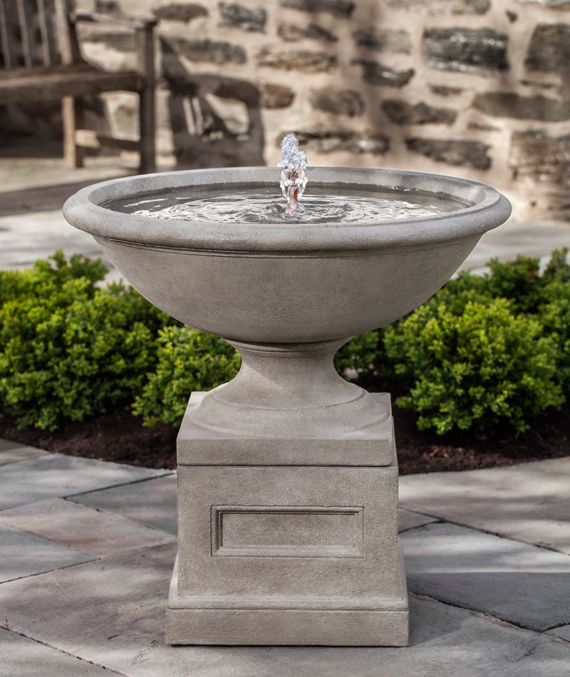 Round basin fountain placed on a square pedestal, pictured in front of a stone wall and low boxwood hedge