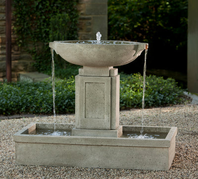 An oval basin pours water from both sides into a rectangular basin, pictures on gravel and in front of a low boxwood hedge