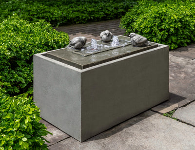 Rectangular fountain with three cast stone birds perched on top plate. Pictured in front of boxwood hedge