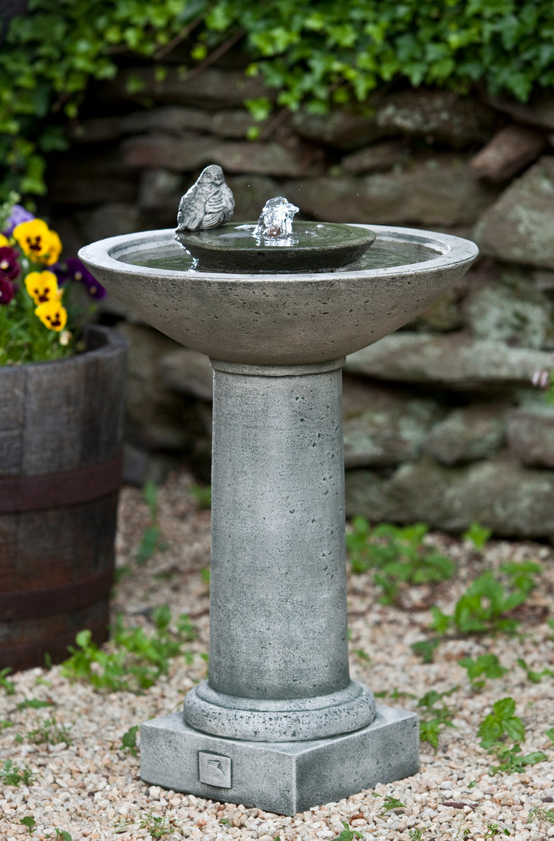 A birdbath style fountain with a small bird perched next to the spout, pictured in front of a stone wall and yellow pansies