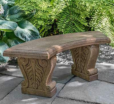 Brown, curved cast stone bench with leaf design on legs, pictured in front of hostas and ferns
