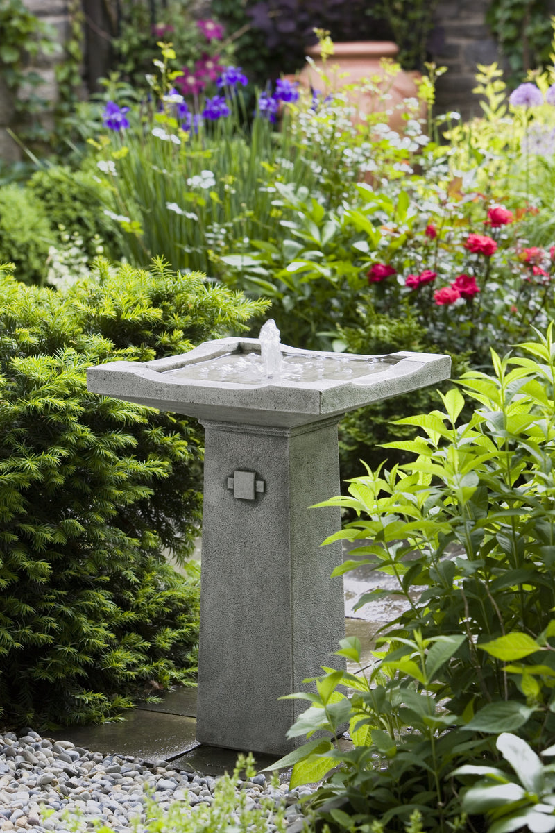 Square basin on a square pedestal with simple decorative medallion, pictured in front of flower garden