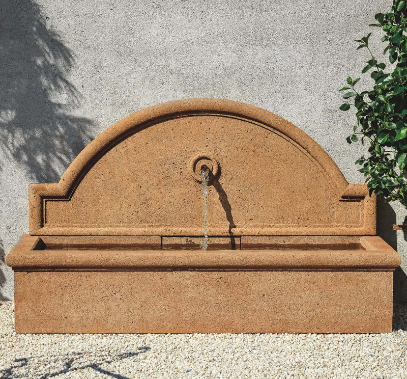 A travertine fountain with a single spout is against a light grey wall.