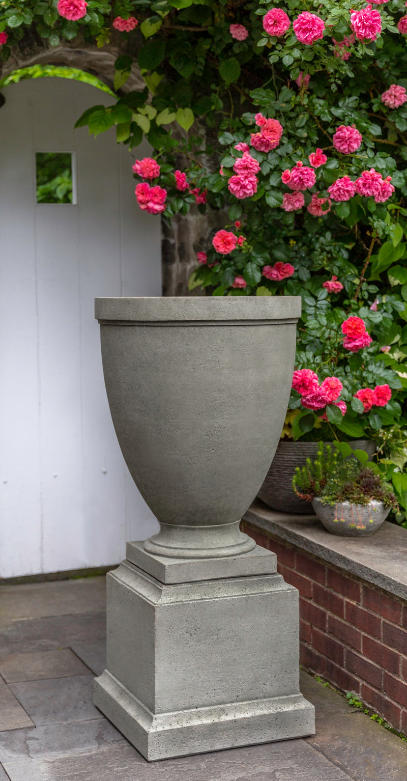 Narrow grey urn pictured on a grey pedestal in front of blooming roses