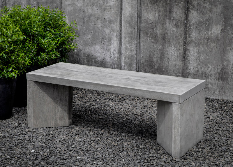 Contemporary grey bench made to look like wood, pictured in front of a grey wall and green plants