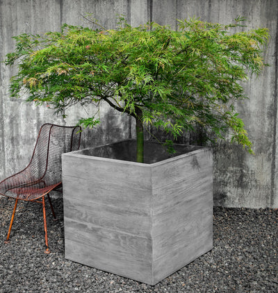 Large square grey container shown planted with a tree and next to a metal chair