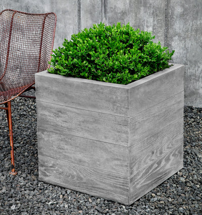 Square cast stone container shown planted with a shrub and next to a metal chair
