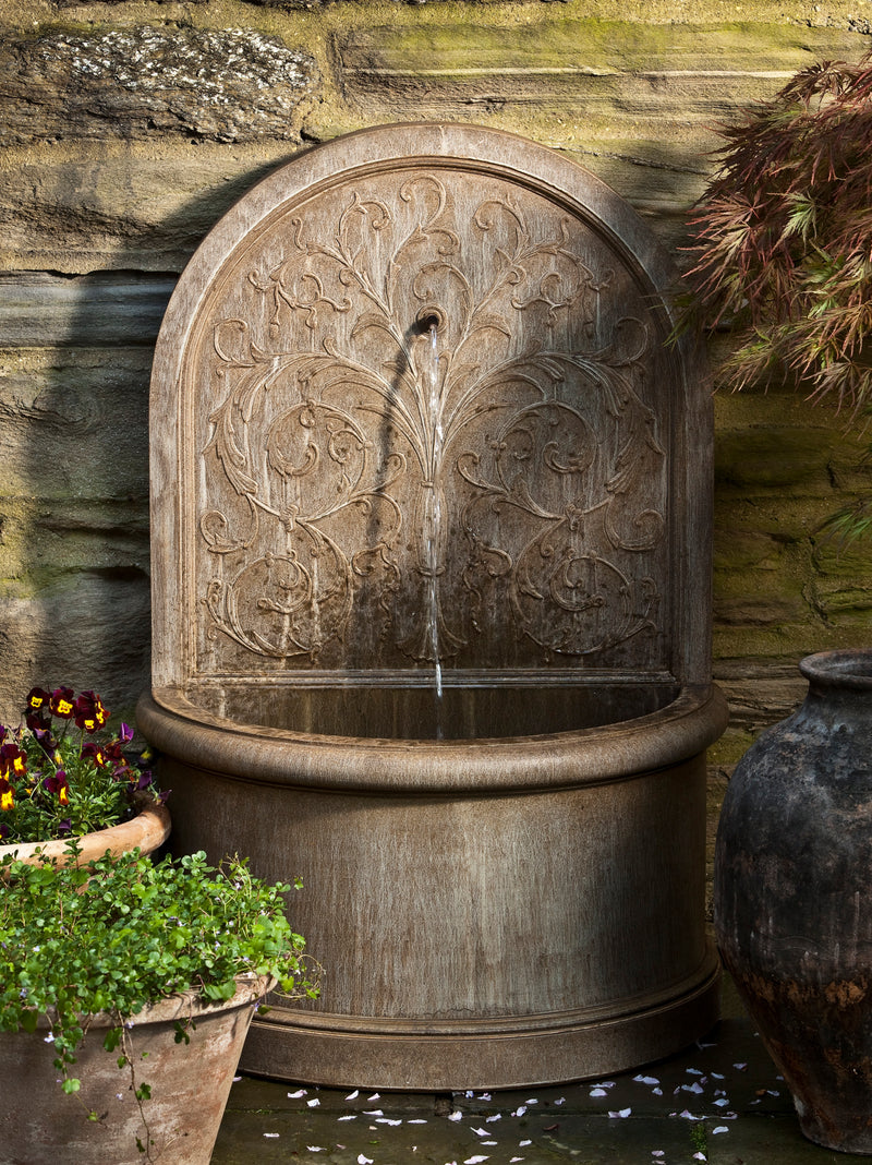 Light brown wall fountain with etched botanical details and water flowing from a spout flanked by planted containers