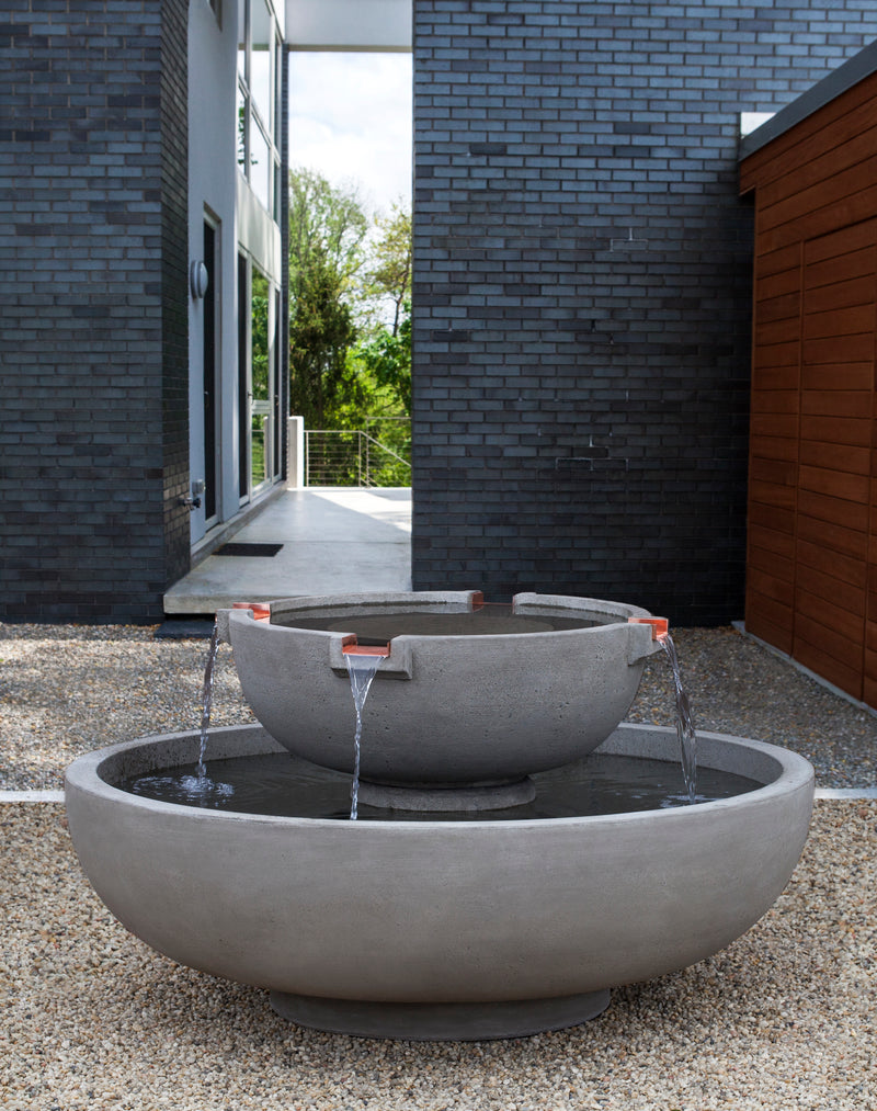 Tiered fountain with two round bowls and four cupper spouts displayed on gravel in front of house