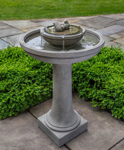 Birdbath fountain in grey finish with sitting birds on top bowl, pictured in front of boxwood hedge
