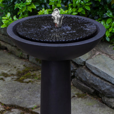 Black birdbath fountain with covered top and water spout in the middle pictured in front of grey stone wall