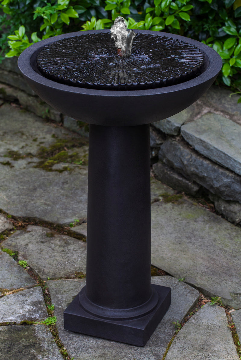 Black fountain with round covered top and water flowing out of copper spout with tall round pedestal pictured on grey stone ground