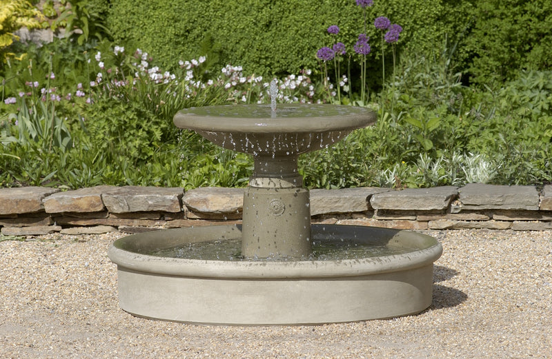 Round in-basin fountain with low top bowl spilling water through center spout pictured in front of rock wall and perennial garden