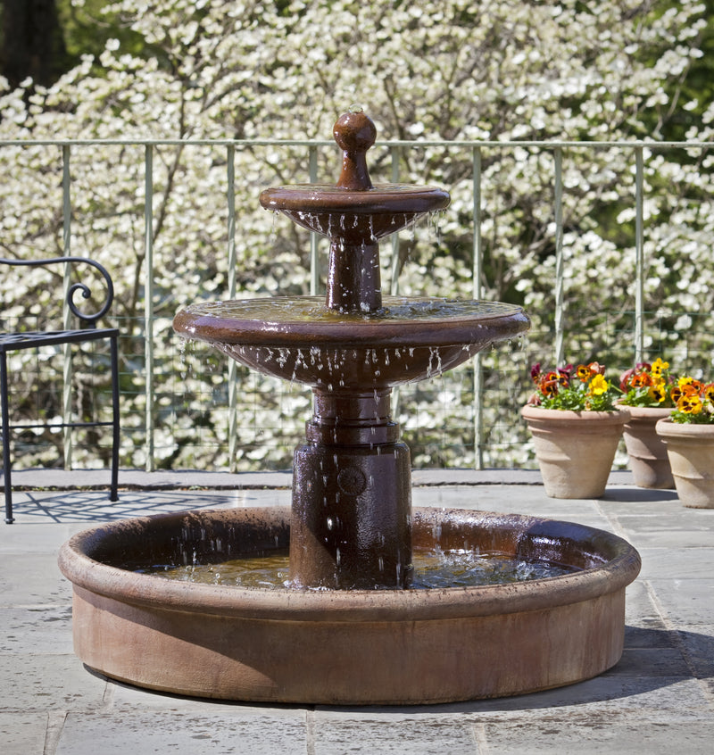 Two-tier brown fountain with water falling into open basin pictured in front of flowering tree