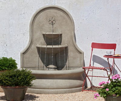Large light brown wall fountain with top medallion and two spilling basins pictured  with red bistro chair and planted plants