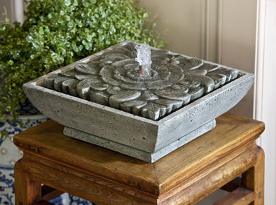 Square tabletop fountain with water spouting from the center pictured on top of wooden stool and greenery in the back