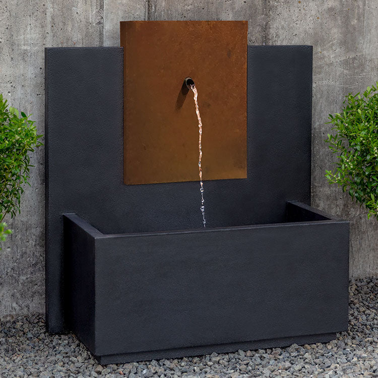 Rectangular wall fountain with copper spout coming out of core ten plaque, pictured in front of concrete wall 