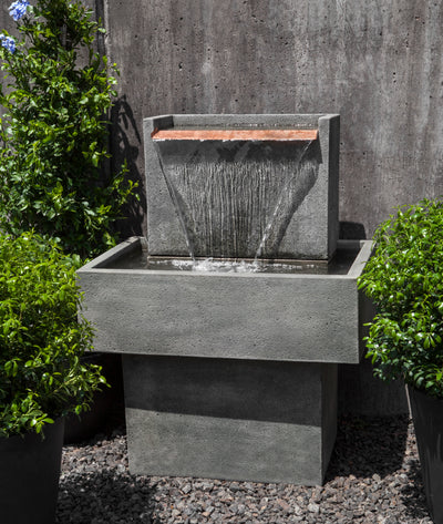 Grey wall fountain with rectangular basin and water falling from large flat copper spout flanked by potted greenery