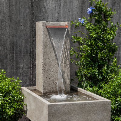 Low rectangular basin with tall narrow back including a flat copper spout with water flowing down pictured in front of wall and flanked by plants
