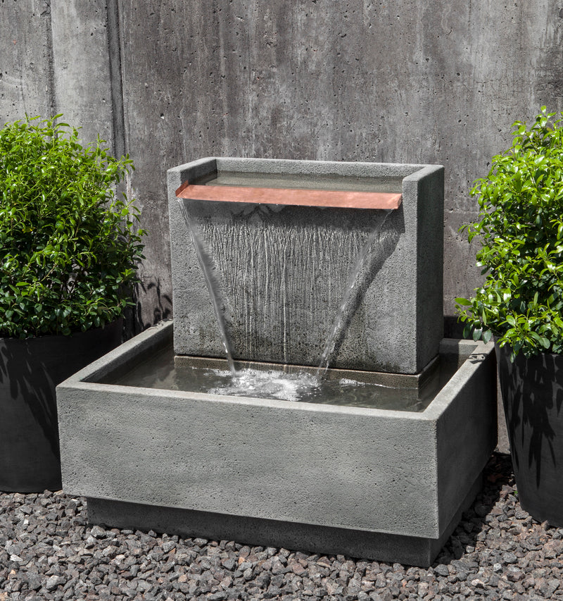 Low rectangular wall fountain with large flat copper spout on top flanked with two potted containers