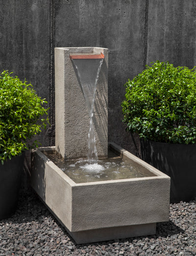 Low rectangular basin filled with water with  tall narrow back and water flowing down from flat copper spout flanked by two potted shrubs