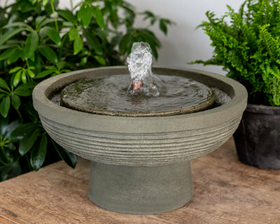 Round grey bowl shaped fountain with covered top and water flowing from copper spout, pictured on top of bench
