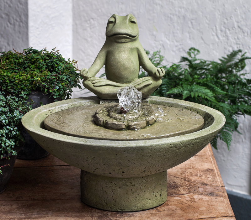 Green frog in a yoga pose on the side of a round fountain pictured  in front of green plants