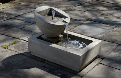 Rectangular basin with an oval shaped bowl  in the back spilling water from a copper spout  and sitting on square stone floor
