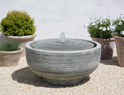 Round fountain with covered top and copper spout pictured on gravel and flanked by terra cotta potted planters