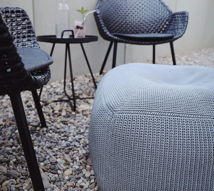 Close up of gray footstool sitting on gravel terrace next to two black chairs
