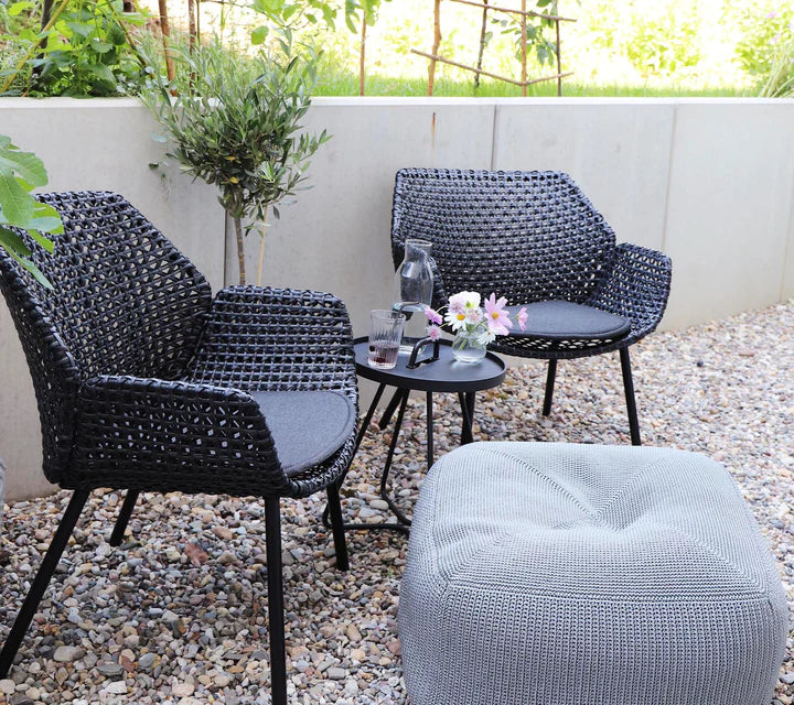 Two black woven armchairs behind a gray footstool on gravel terrace