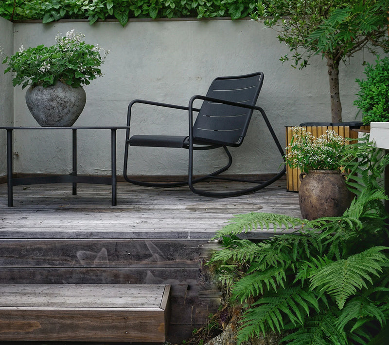 Black contemporary rocking chair on top of raised wooden deck and next to matching coffee table with fern in front